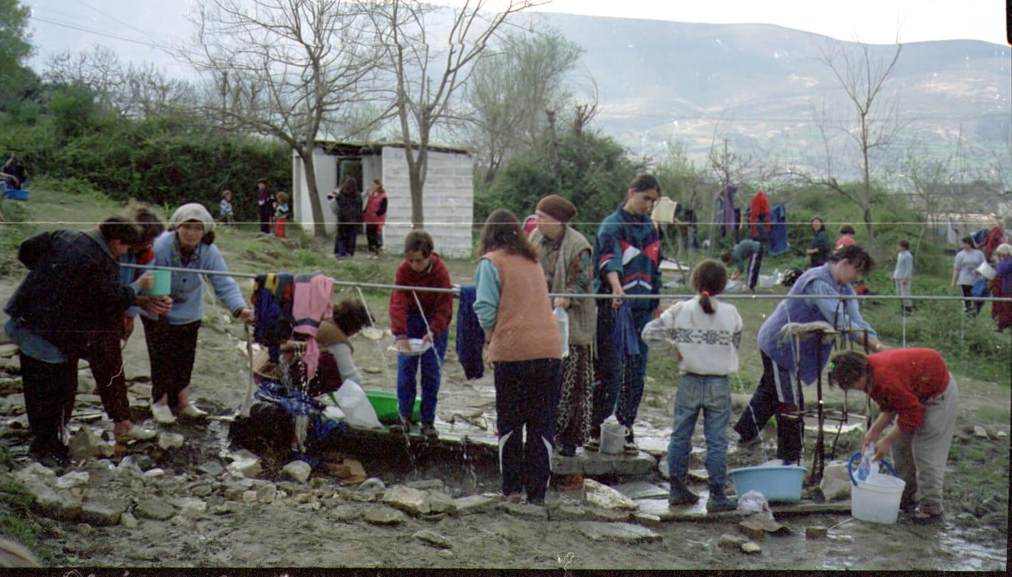 Women and children wash clothes and fetch water at an outdoor water point; buildings and hills behind.