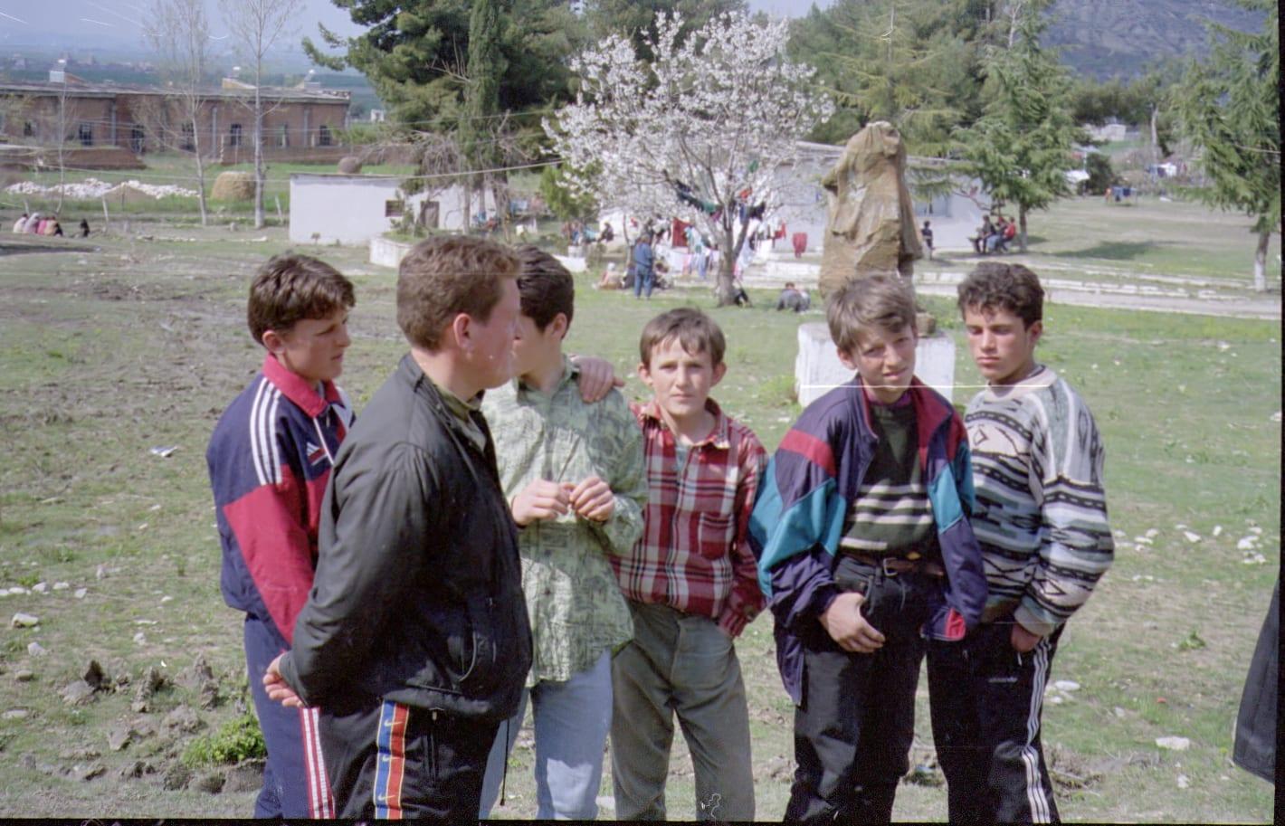 Six boys stand together on a grassy field; tents and a blossoming tree in the background.