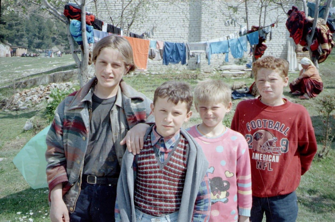 Four children pose together; behind them laundry on a line and a wall.