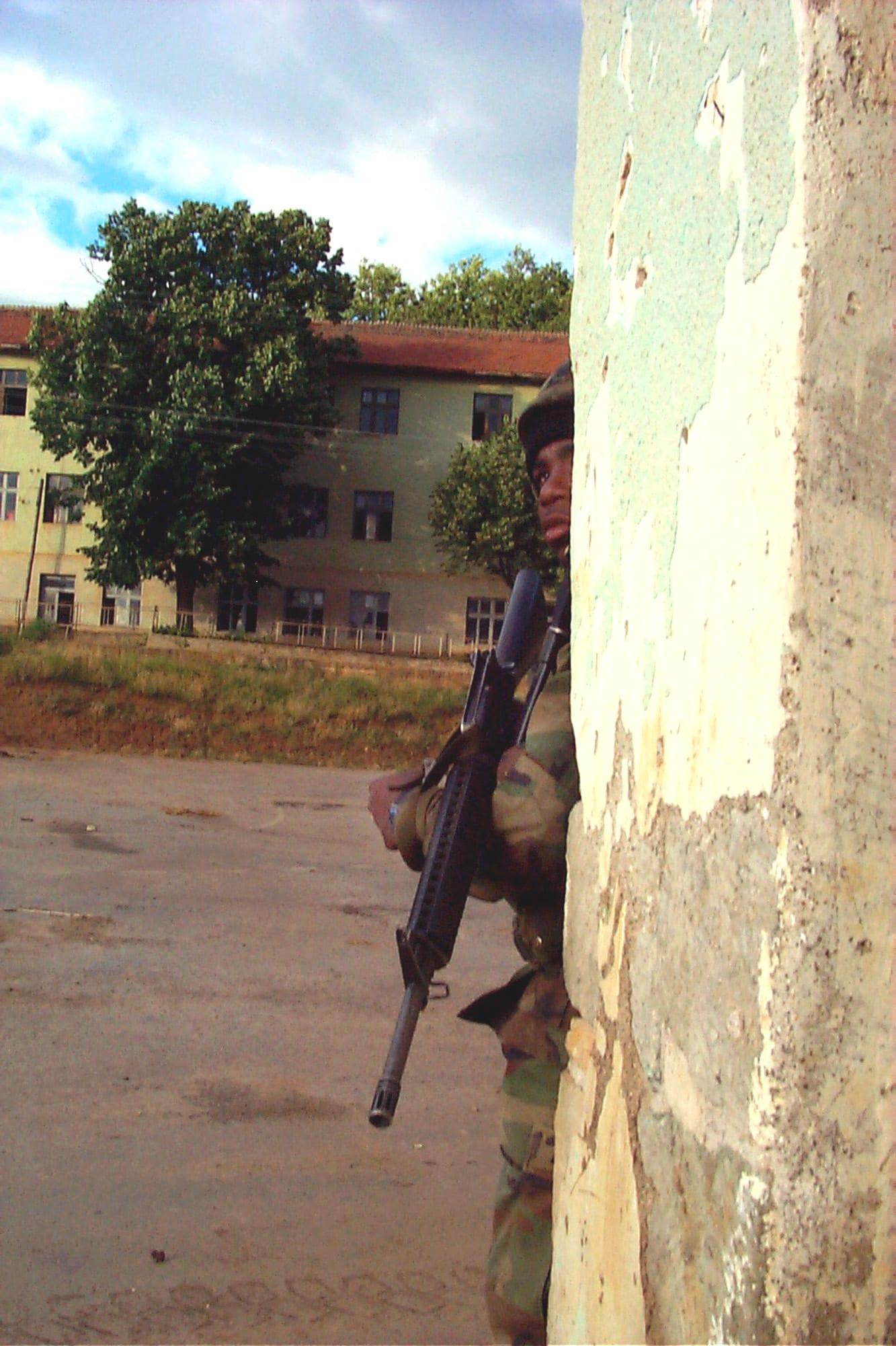 A soldier with a rifle sheltering behind a concrete pillar, a building in the background.