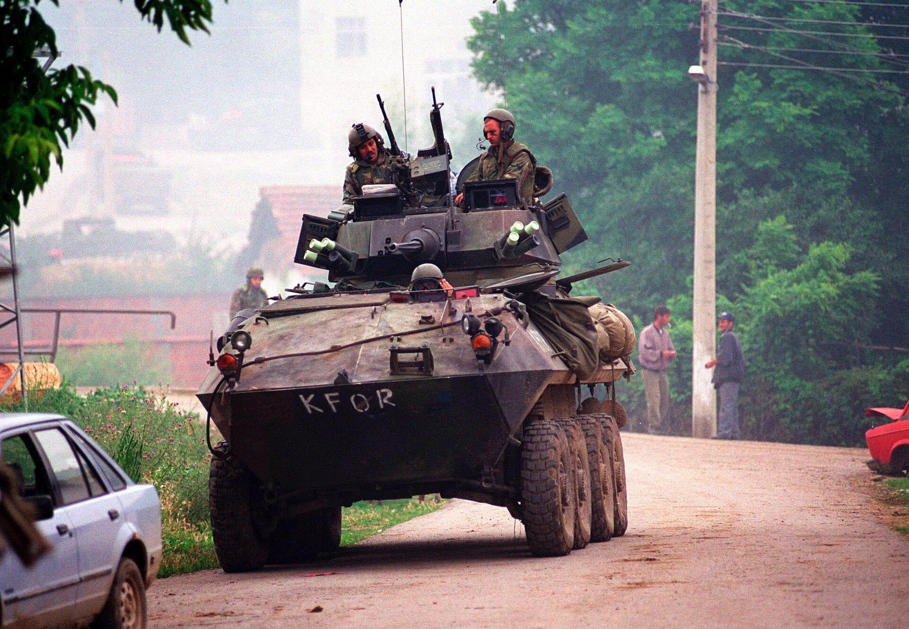 Armoured vehicle marked KFOR on a village road, with soldiers in the turret.