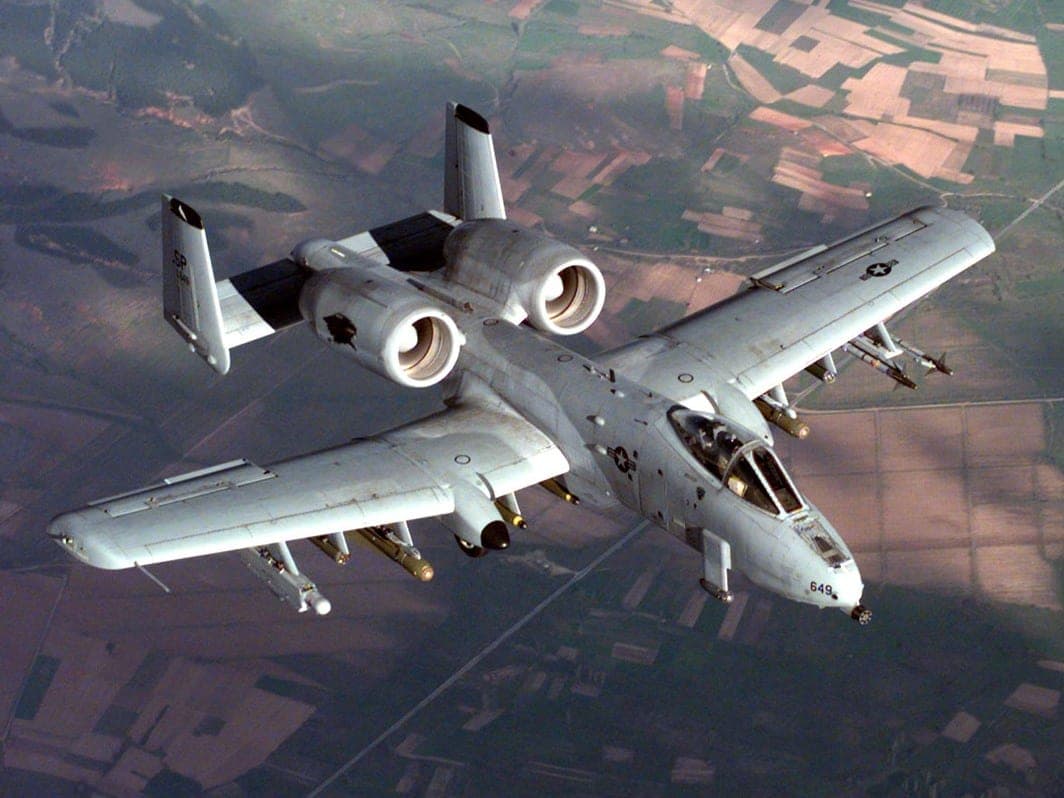 A grey attack aircraft with two engines above the fuselage flying over farmland.