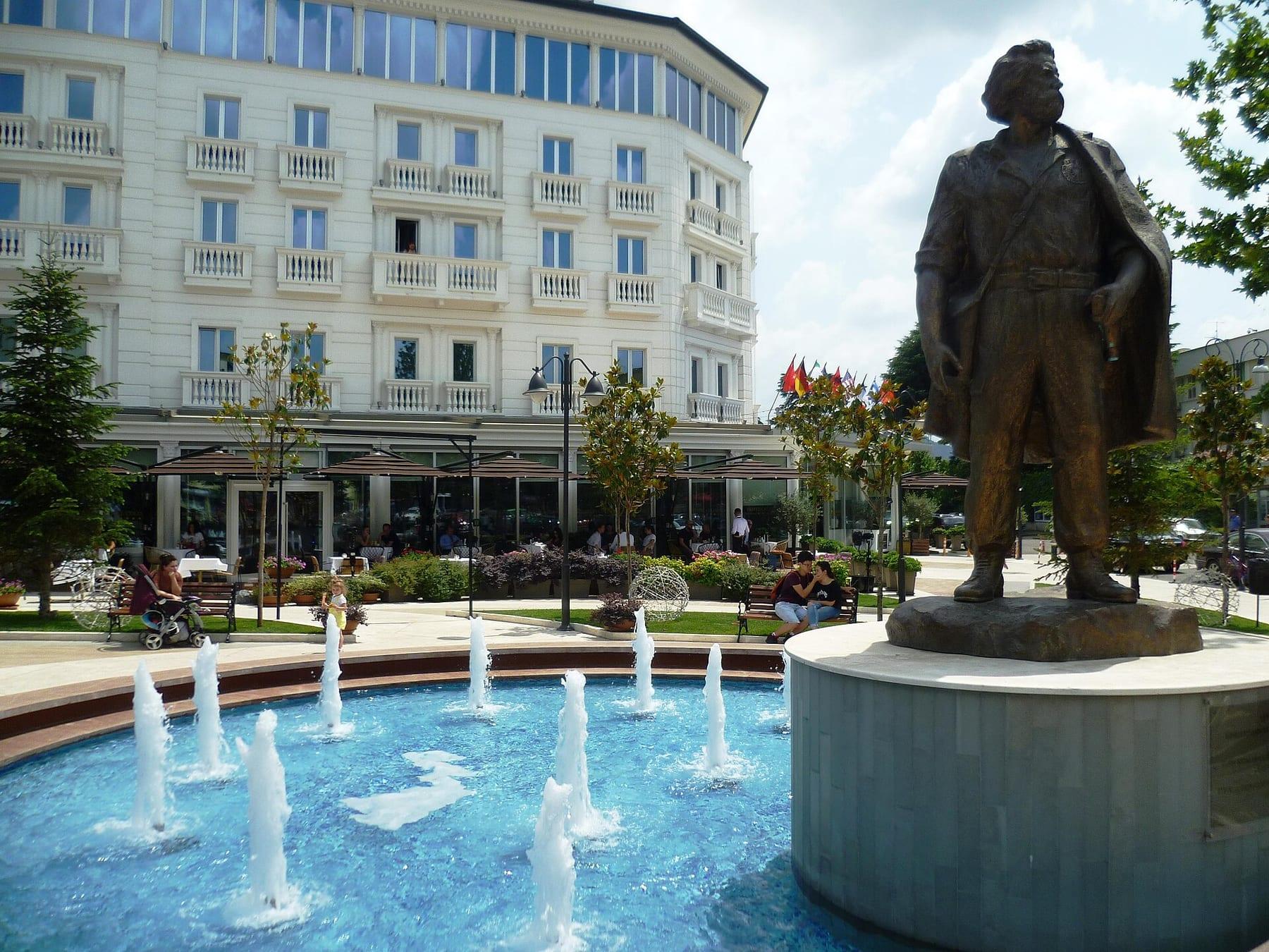 Bronze statue of a standing man beside a round fountain, with buildings behind.