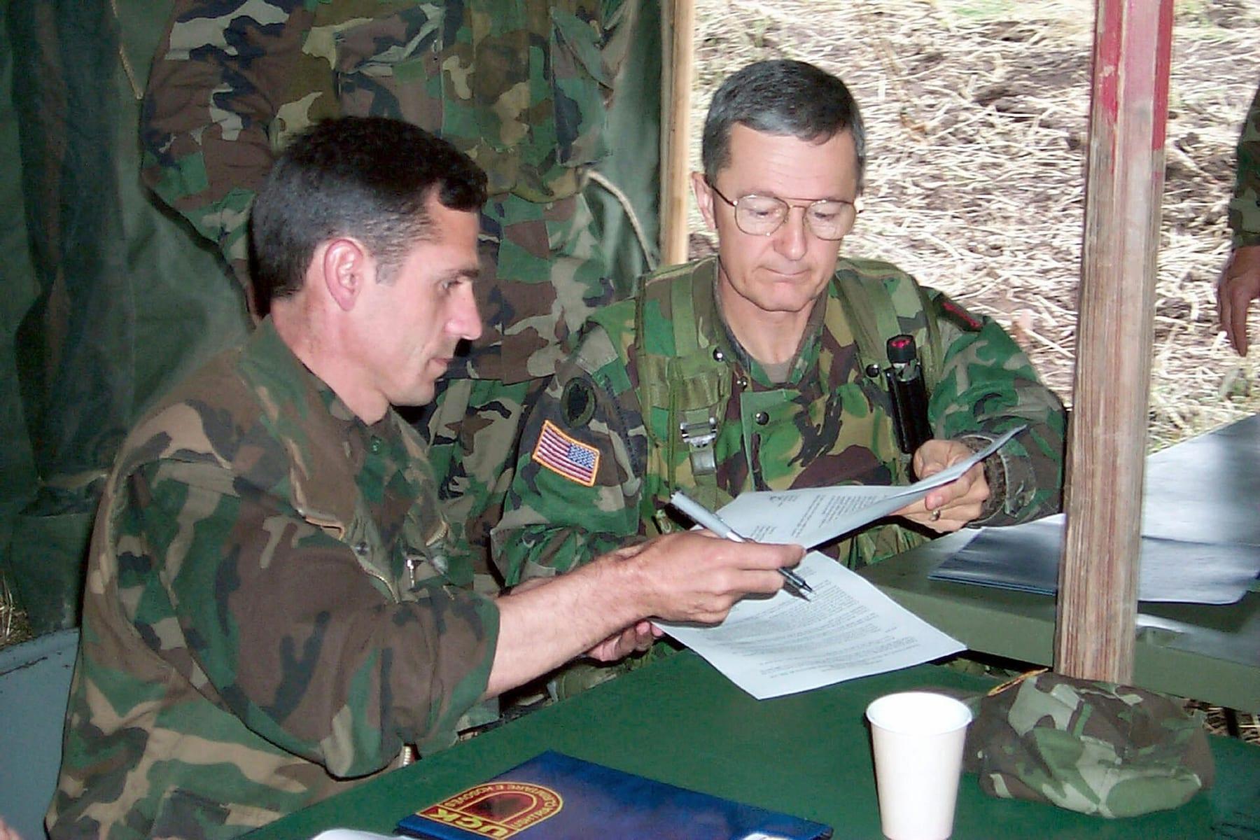 Two officers in camouflage uniforms seated at a table under a tent exchanging papers.