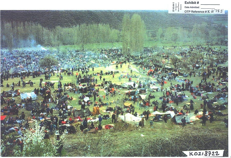 A field crowded with people, plastic sheeting and smoke from fires, seen from a distance