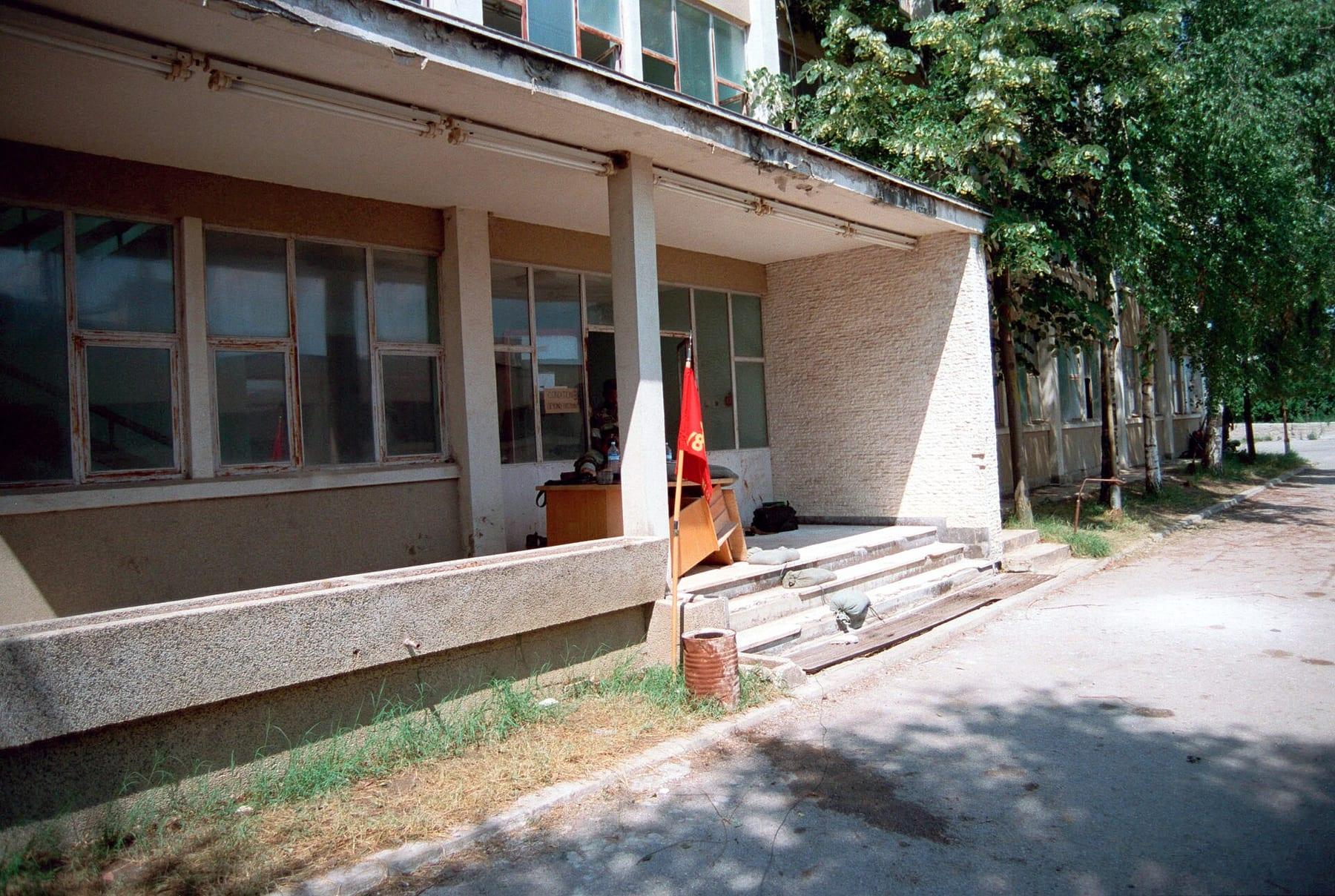 Single-storey building with a concrete canopy and steps on a tree-lined street.
