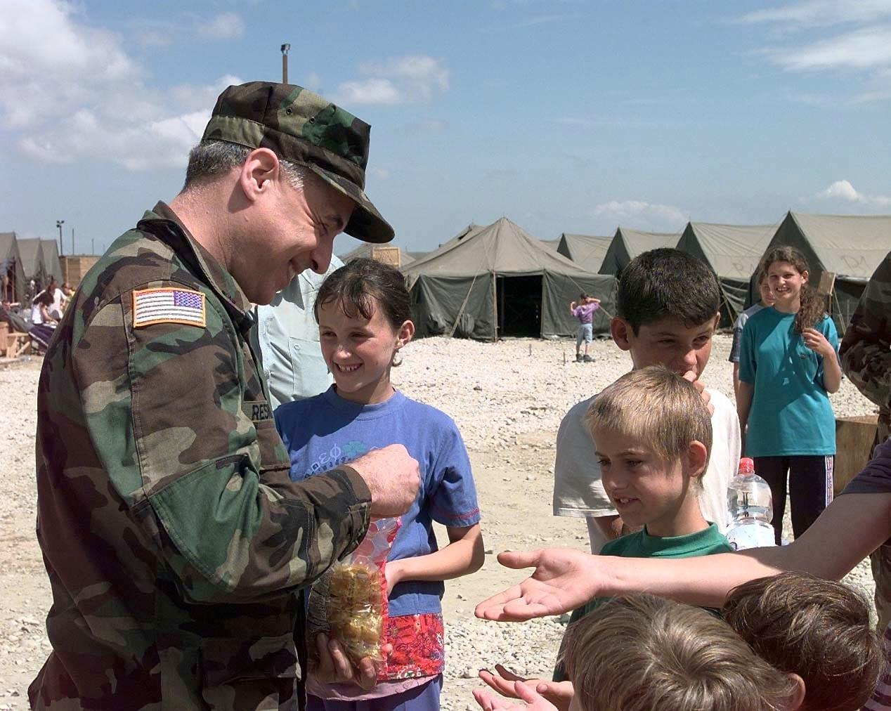 A US serviceman hands out sweets to smiling children; military tents behind.
