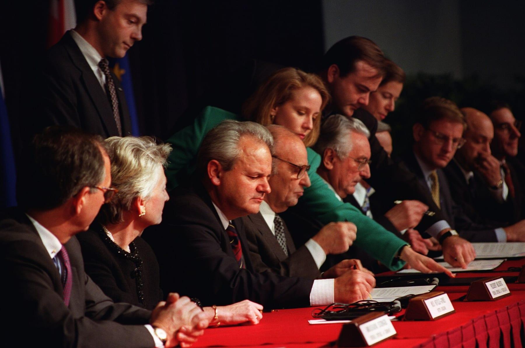 Three men seated at a red-covered table signing documents, surrounded by diplomats.