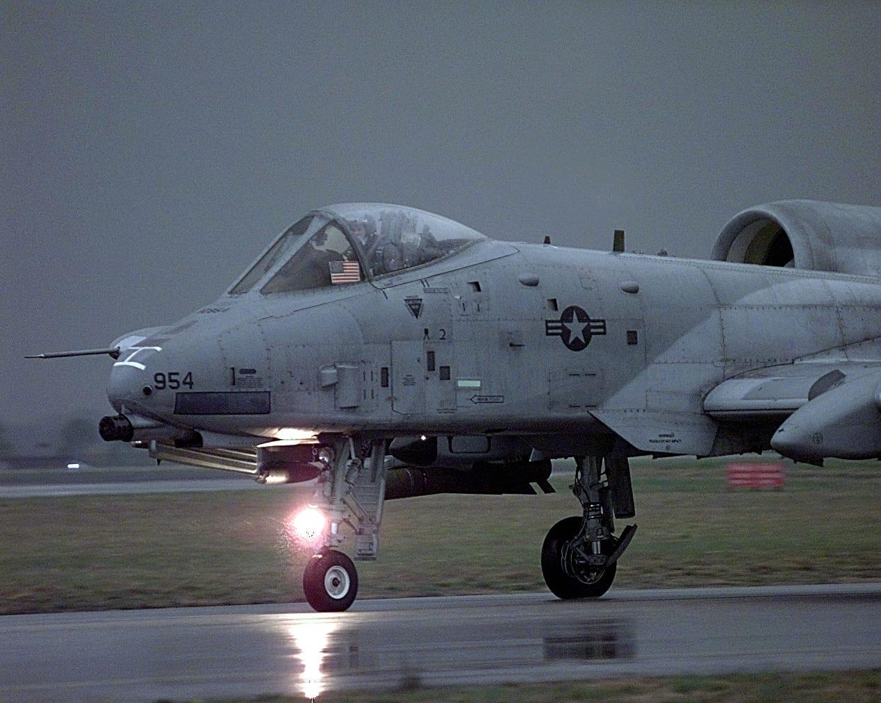 Grey attack aircraft with its light on, on a wet taxiway.