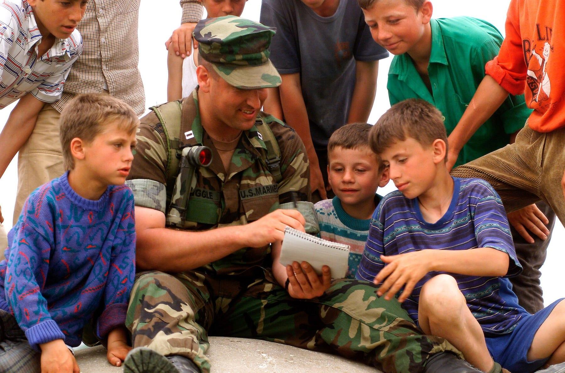 A uniformed soldier sitting among smiling boys looking at a notepad.