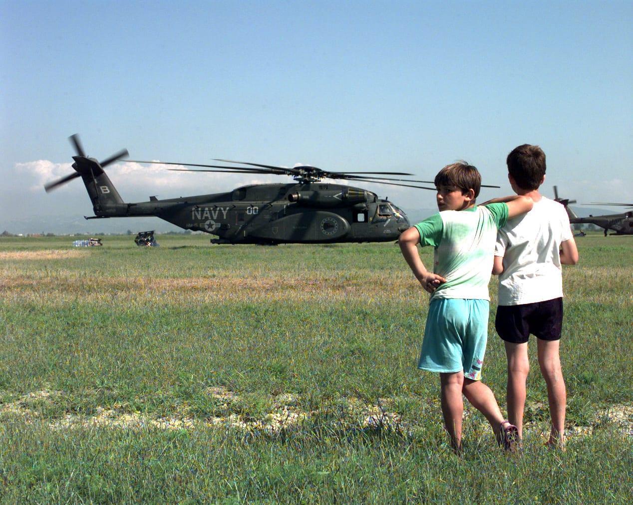 Two boys seen from behind, arms around each other, watching a helicopter in a field.