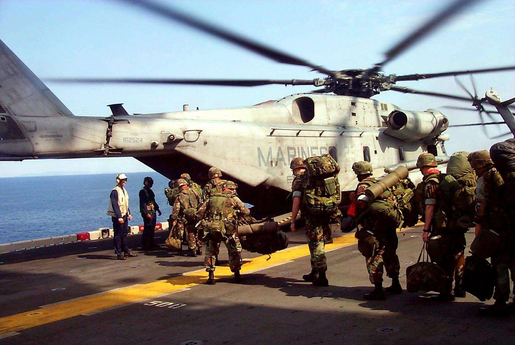 Marines with rucksacks boarding a large helicopter on a ship's deck, the sea behind.