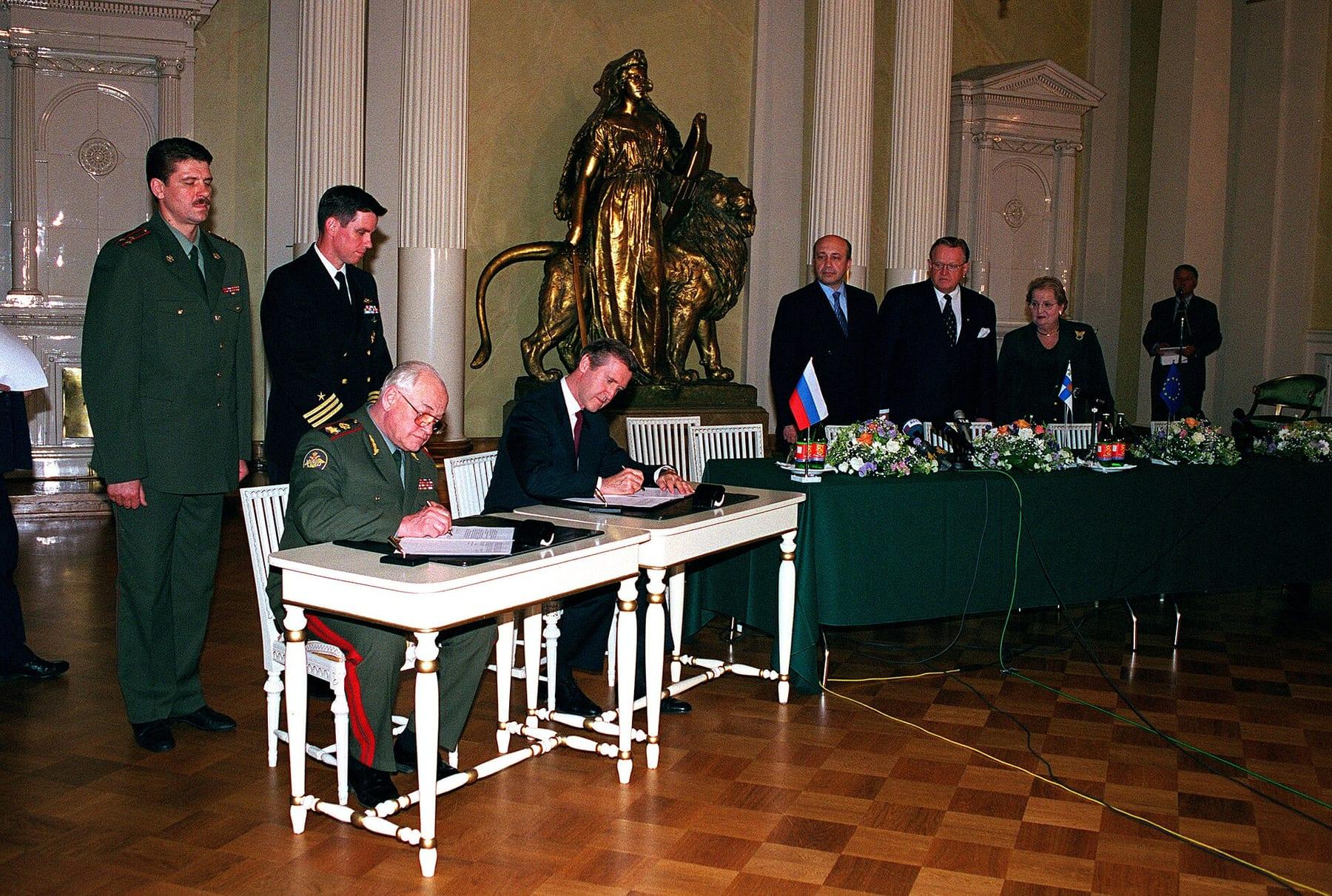 Two men seated at a white table signing documents in a ceremonial hall, officers and diplomats behind them.