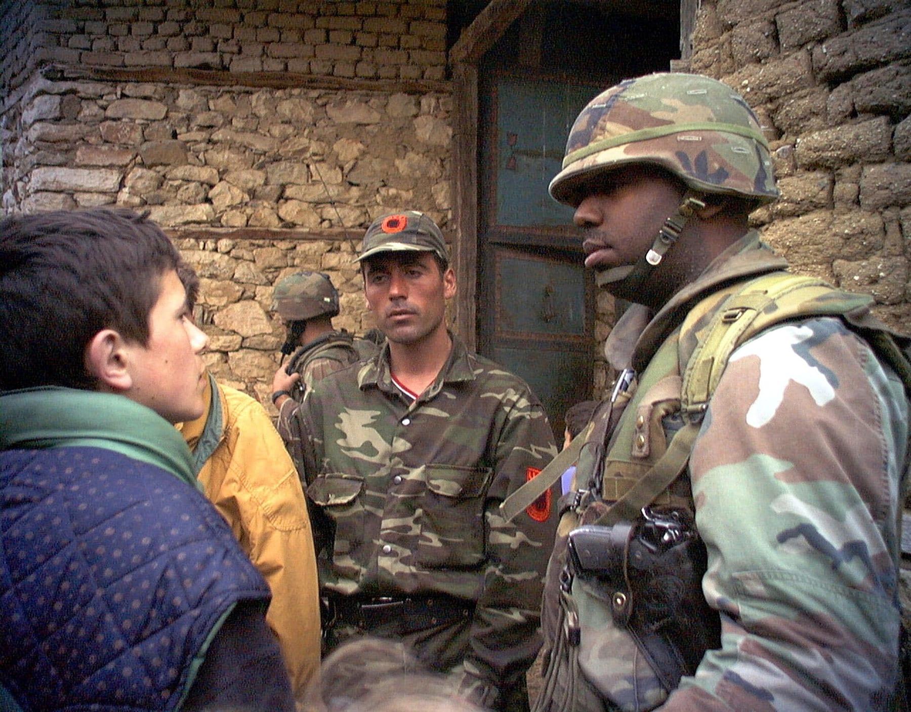 An American soldier in a helmet facing a man in uniform and a cap with a UÇK badge, before a stone wall.