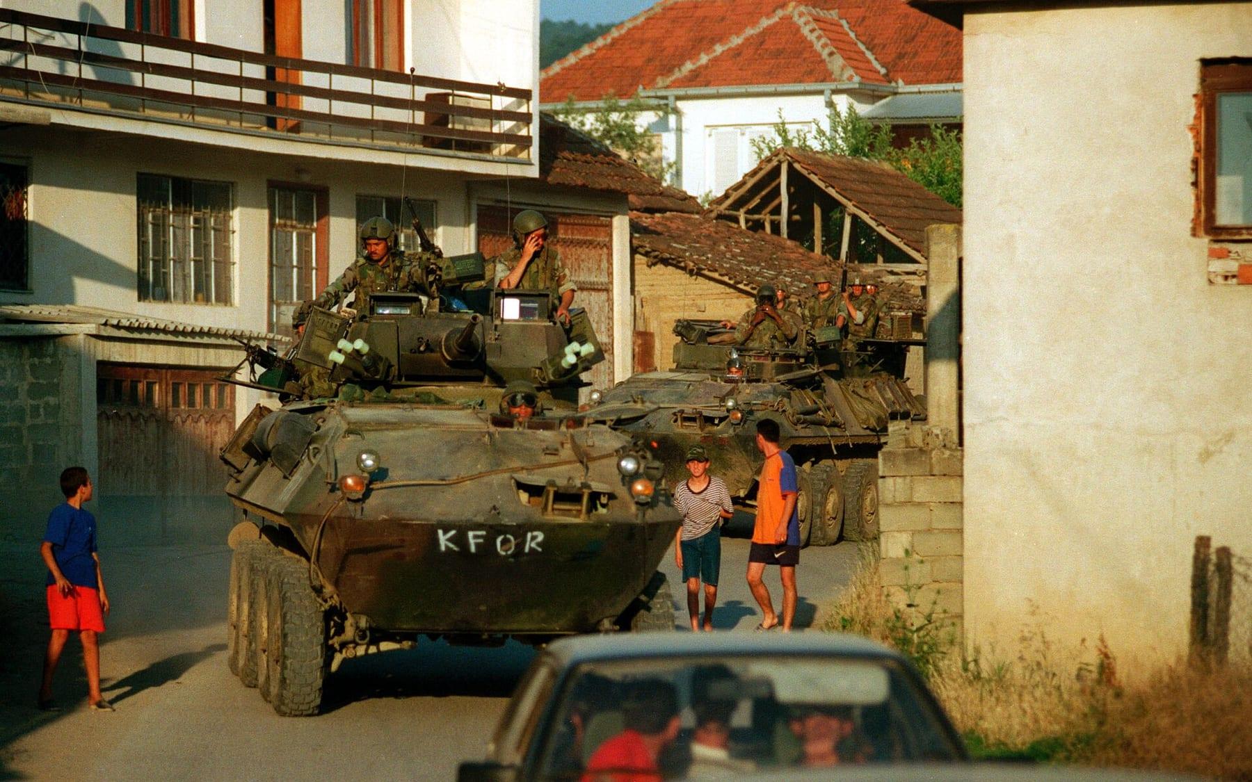 An armoured vehicle marked KFOR passing through a narrow village street as children look on.