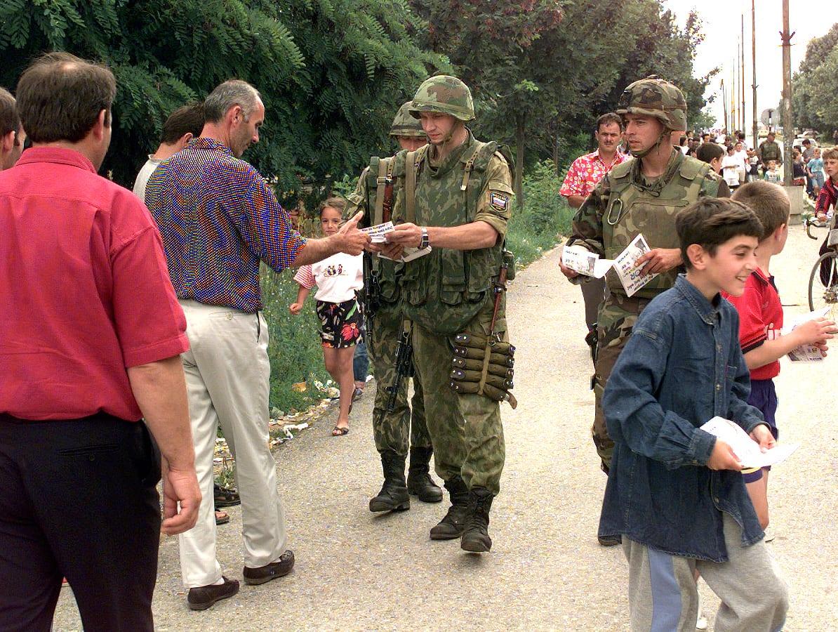 Helmeted soldiers handing leaflets to residents and children on a road.