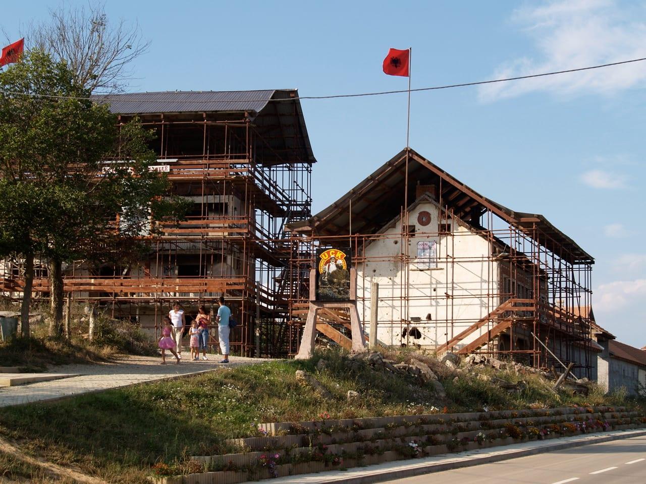 Two damaged houses surrounded by scaffolding with an Albanian flag above.