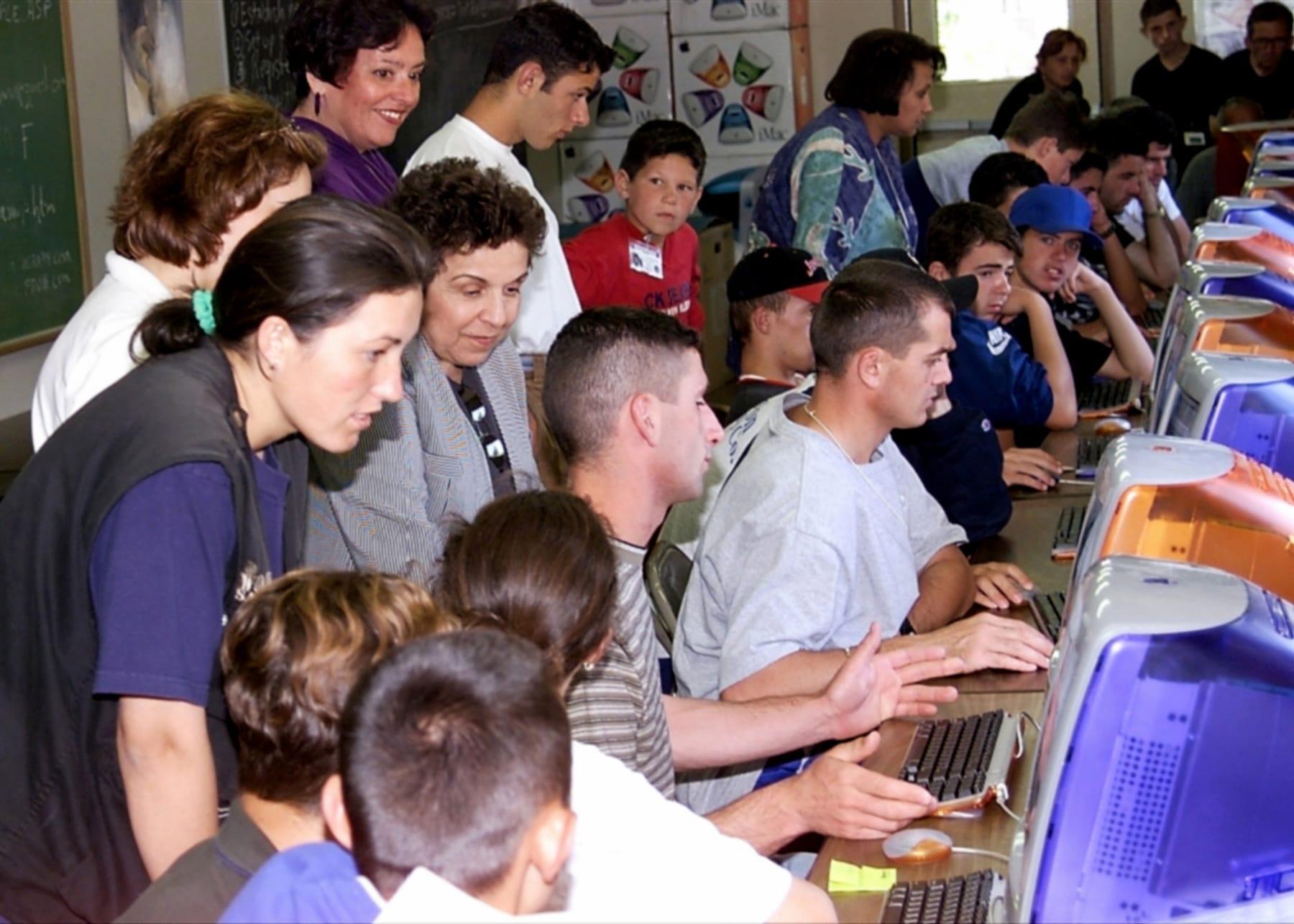 A group of young people and women leaning over iMac computers; a woman in a grey jacket looks on.