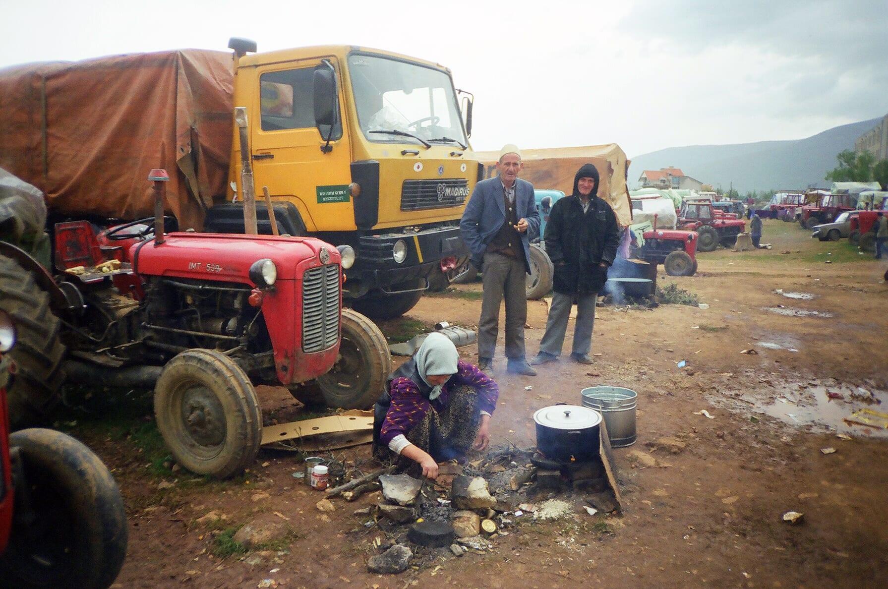 A woman cooks over an open fire beside a red tractor; two men stand in front of a tarpaulin-covered lorry.