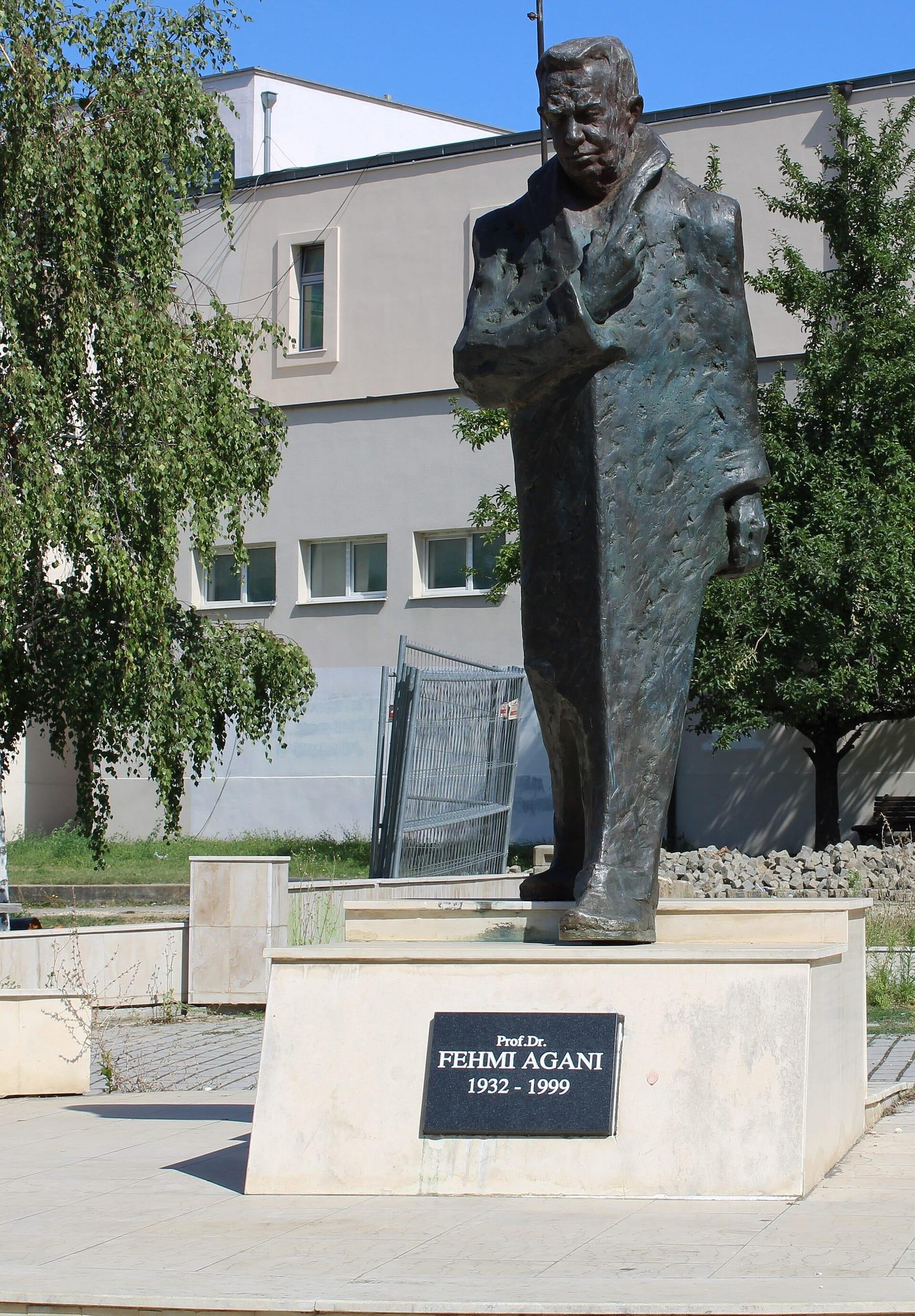 Bronze statue of a standing man on a plinth inscribed “Fehmi Agani”, in front of a building.