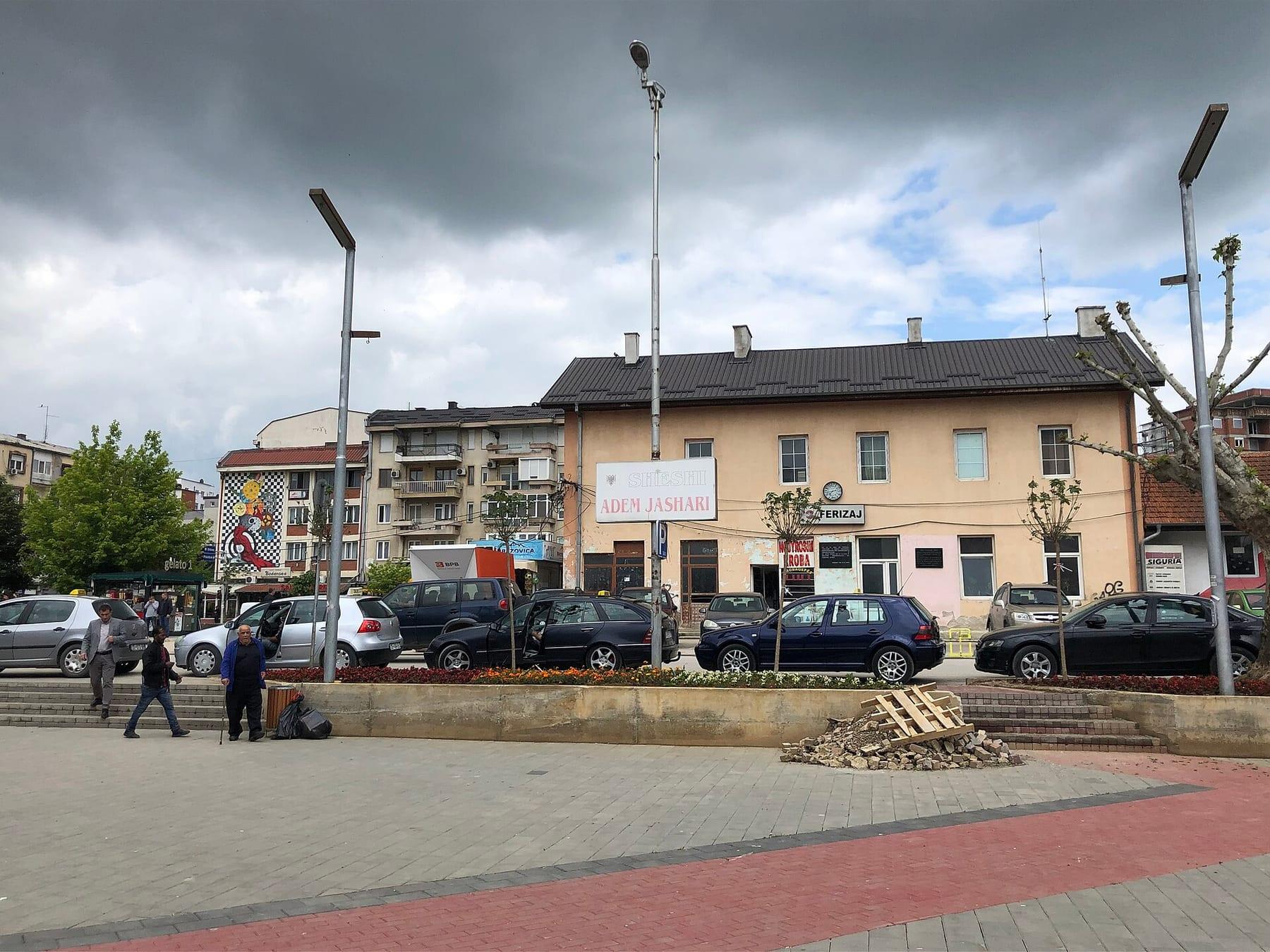 Square with parked cars in front of the yellow railway-station building.