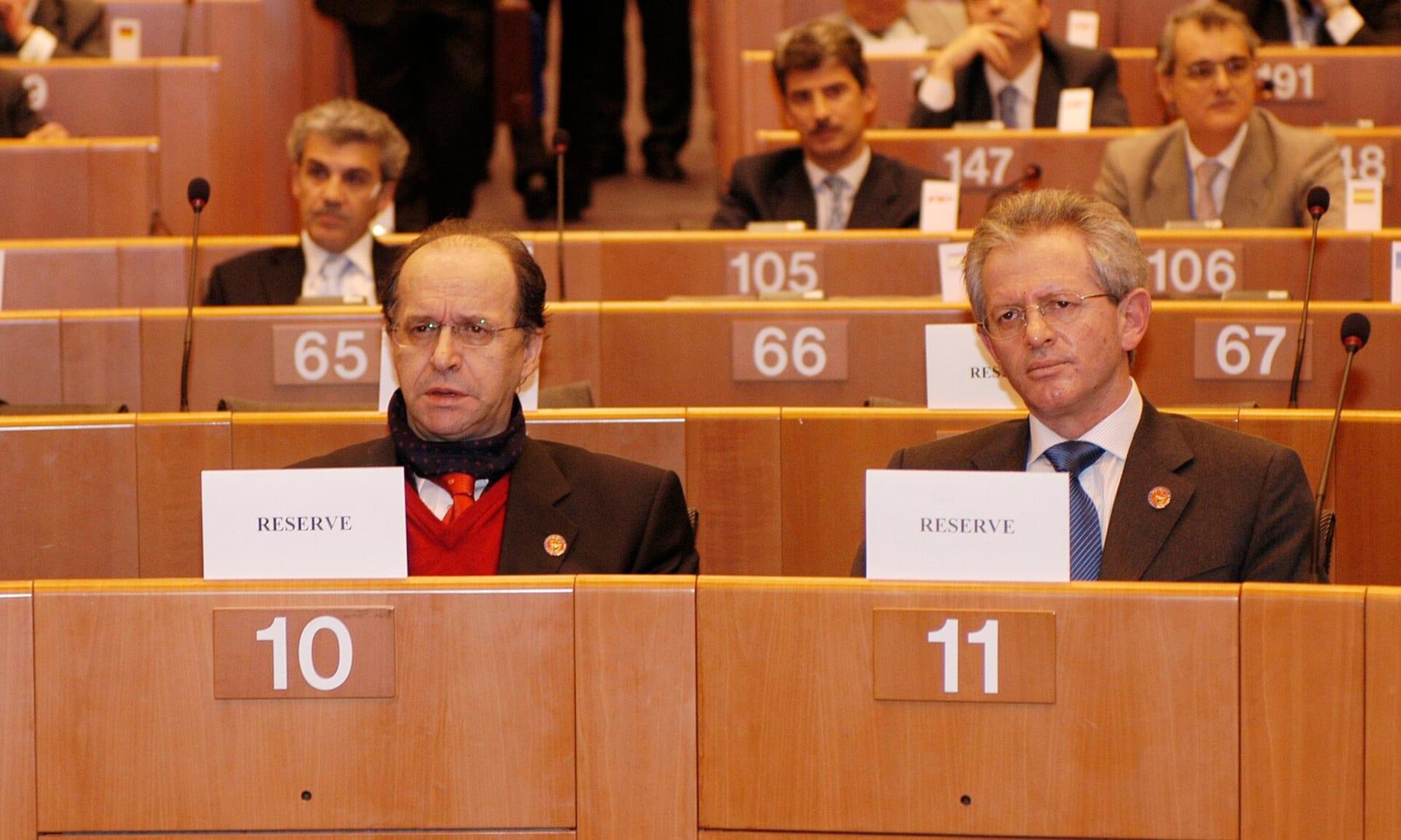 Two men seated in a parliamentary chamber behind “Reserve” signs; one wears a scarf around his neck.
