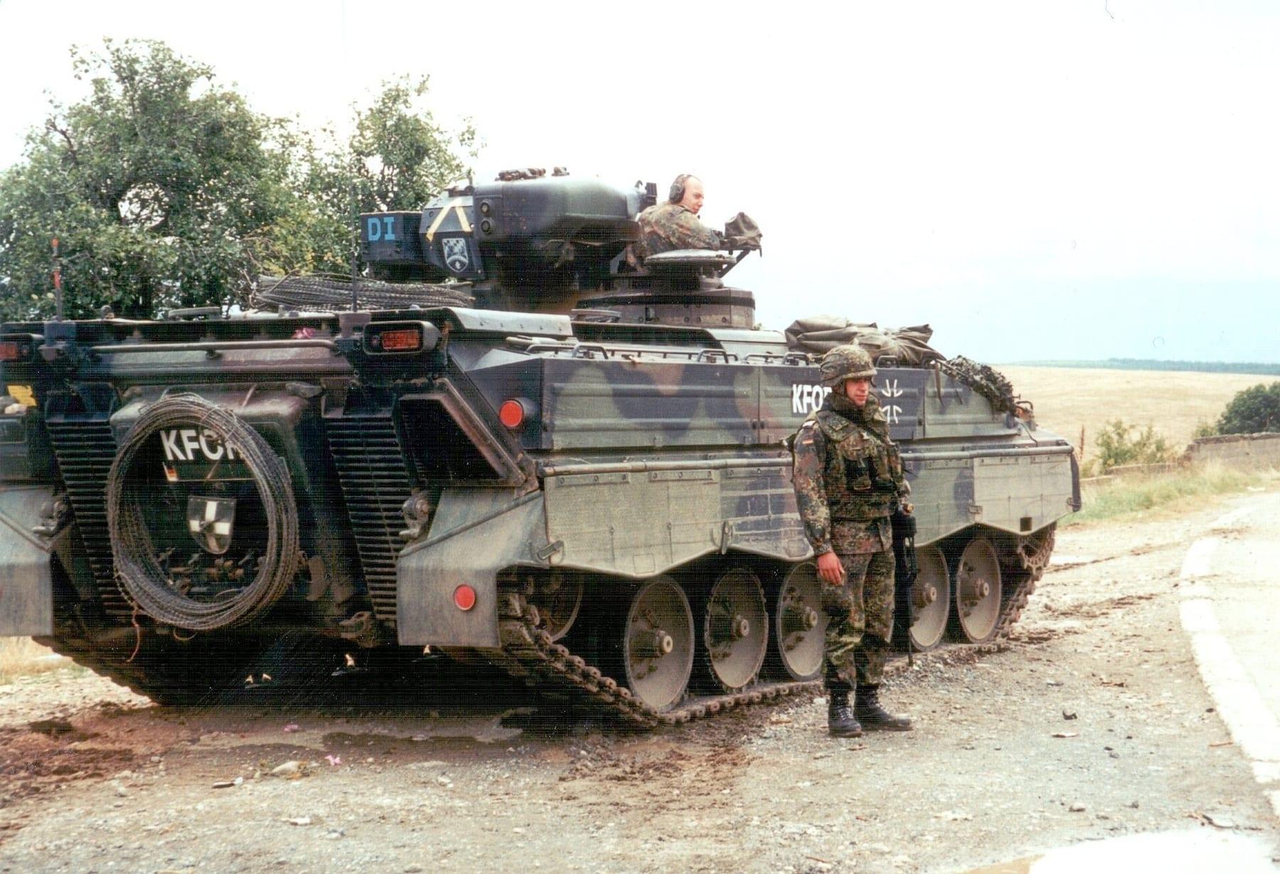 Tracked armoured vehicle marked KFOR with a soldier standing beside it.