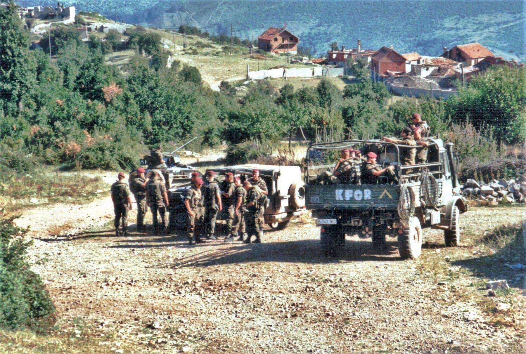 Soldiers beside a truck marked KFOR on a dirt road, with a hillside village behind.