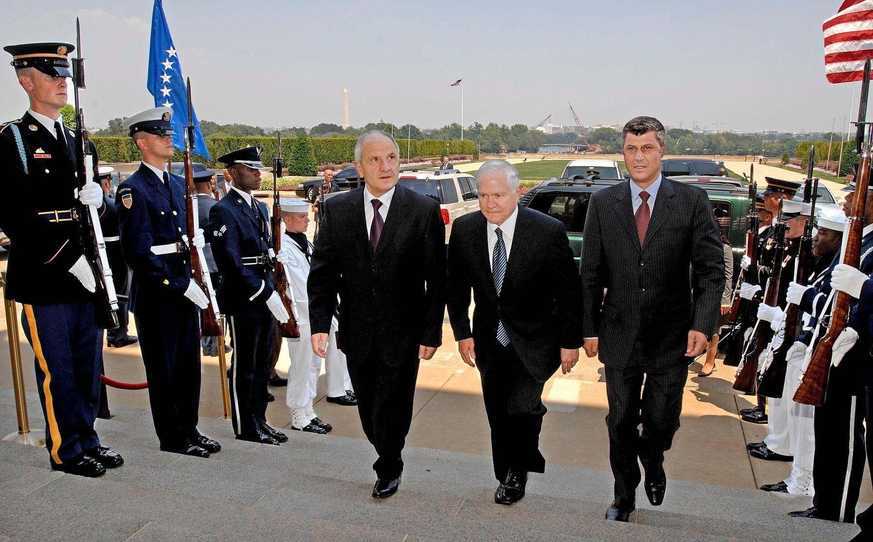 Three men in suits walk through a military honour cordon with flags.