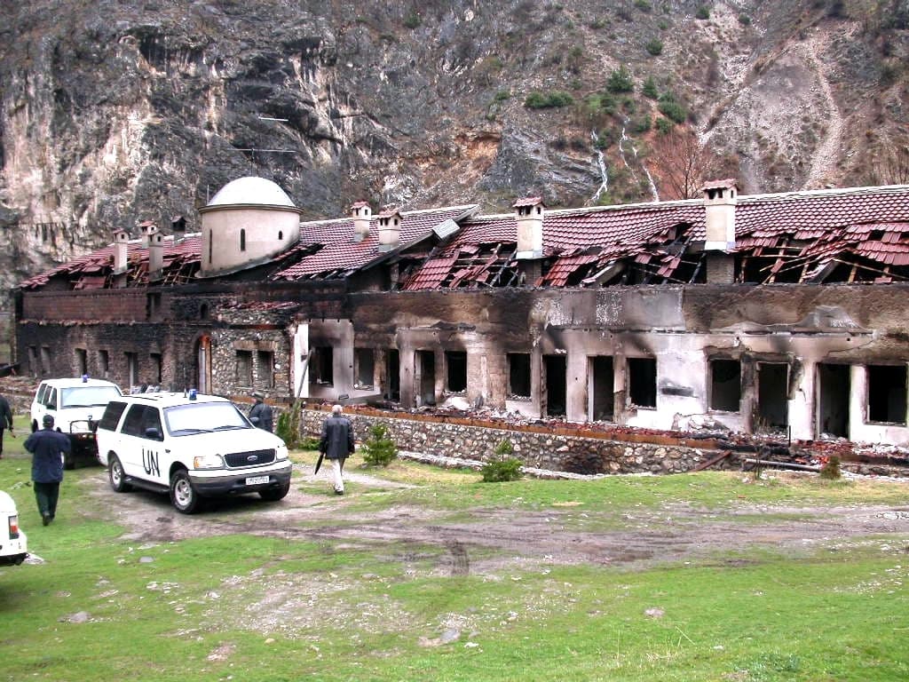 Long burnt-out monastery building with collapsed roof; white UN vehicles in front.