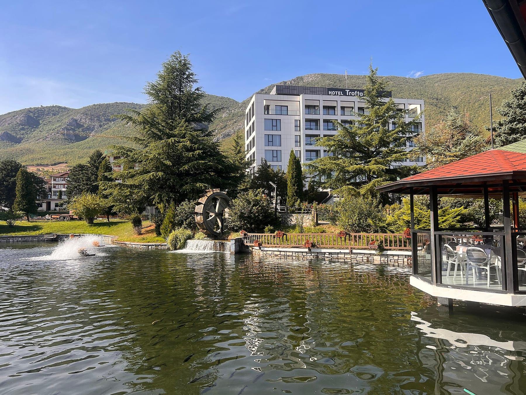 Pond with trees, a wooden gazebo and a hotel, with the Istog mountains behind.