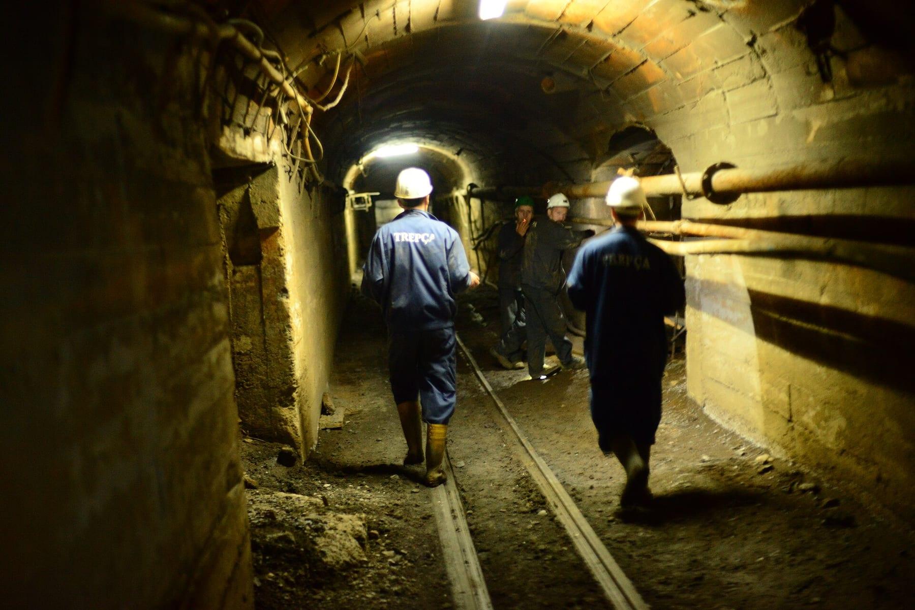 Miners in helmets walk along rails in a dimly lit mine tunnel.