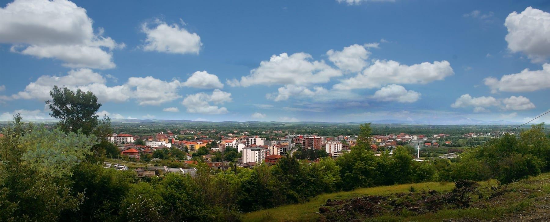Panorama of the town on the plain seen from a green hillside under a cloudy sky.