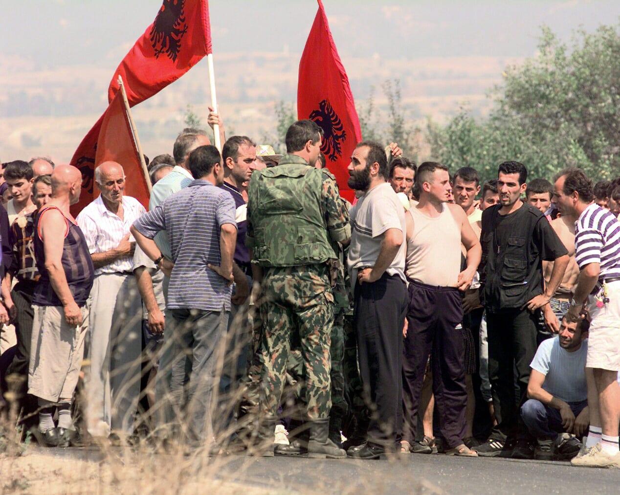 A soldier in camouflage, back to the camera, faces a group of men holding red Albanian flags.