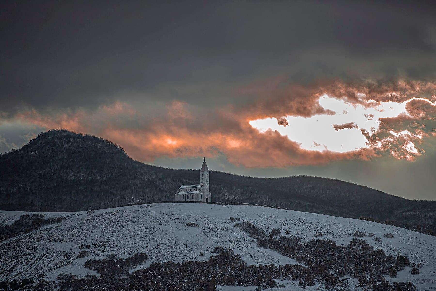 Small white church on a snowy hill under dark clouds at sunset.