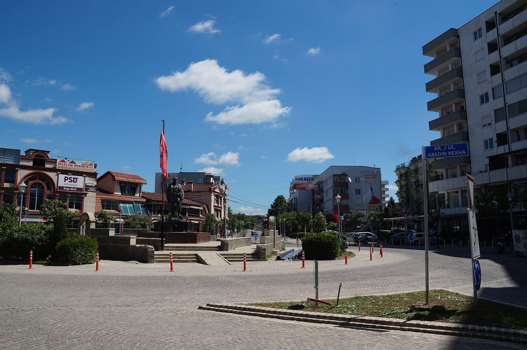 Wide square with a monument, flags and surrounding buildings.