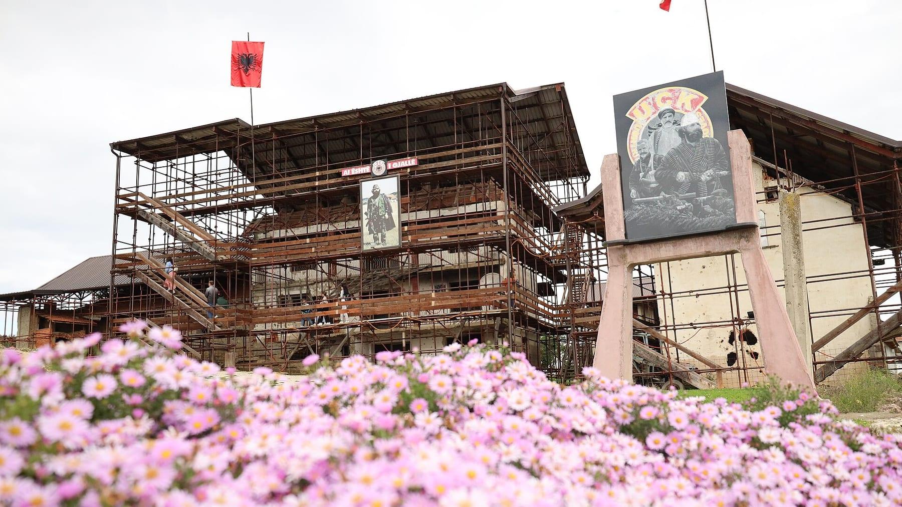 Damaged house enclosed in protective scaffolding, with an Albanian flag and a portrait panel; flowers in the foreground.