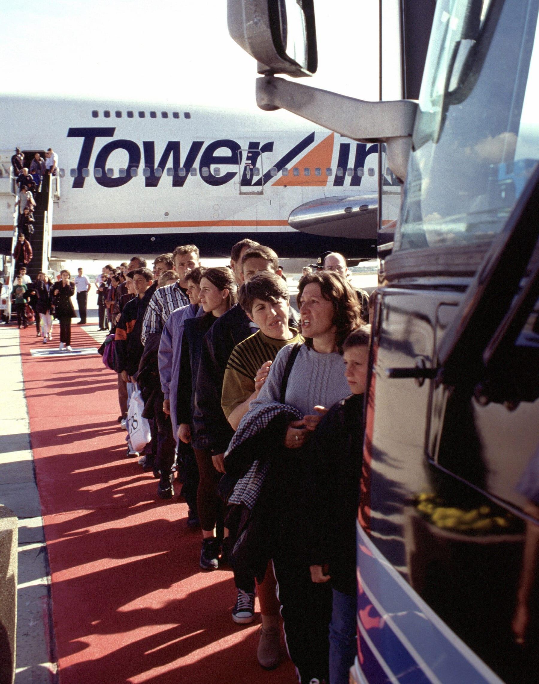 Line of people with bags beside a bus; behind them an aircraft marked Tower Air.