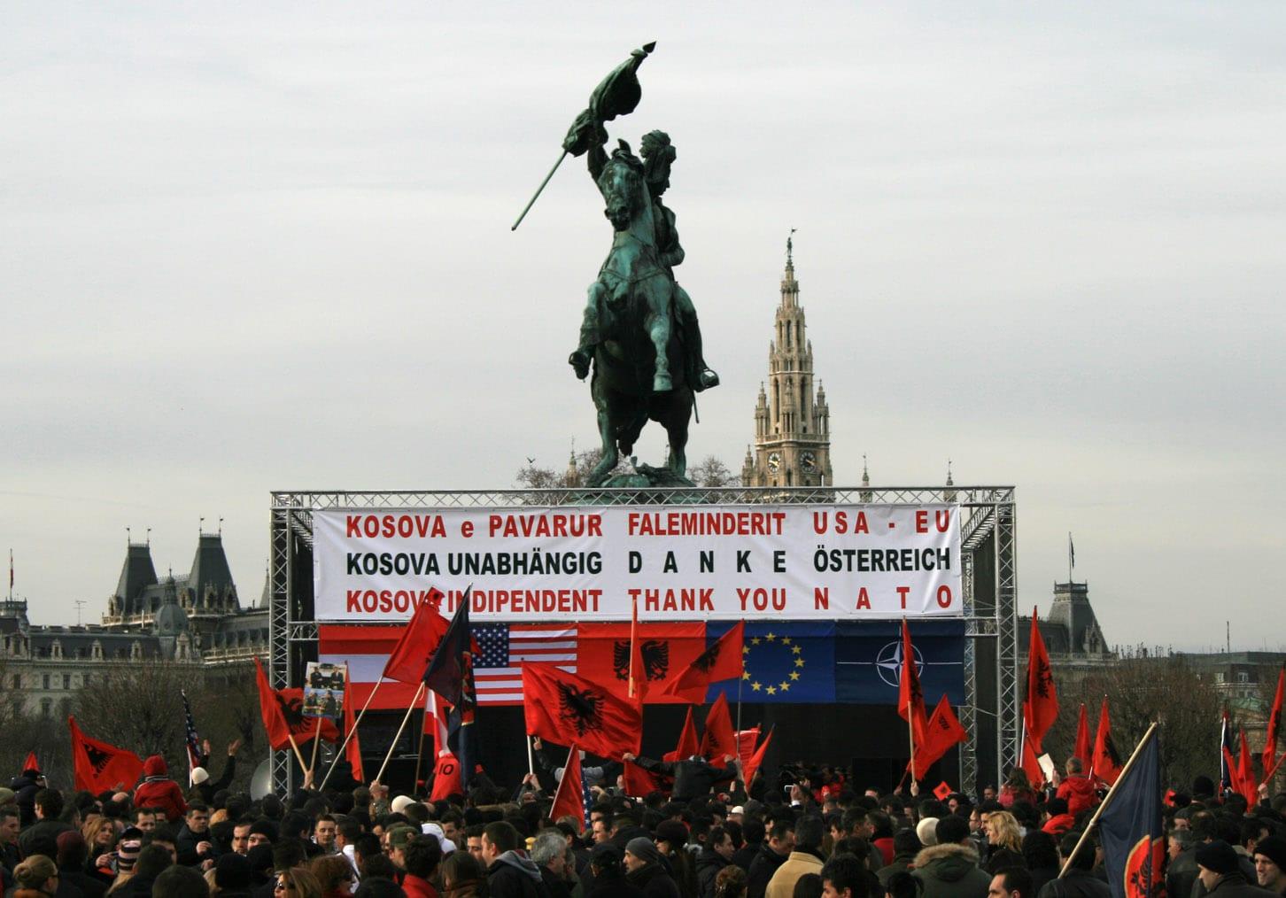 A crowd with Albanian flags before a stage with thank-you banners, beneath an equestrian statue.