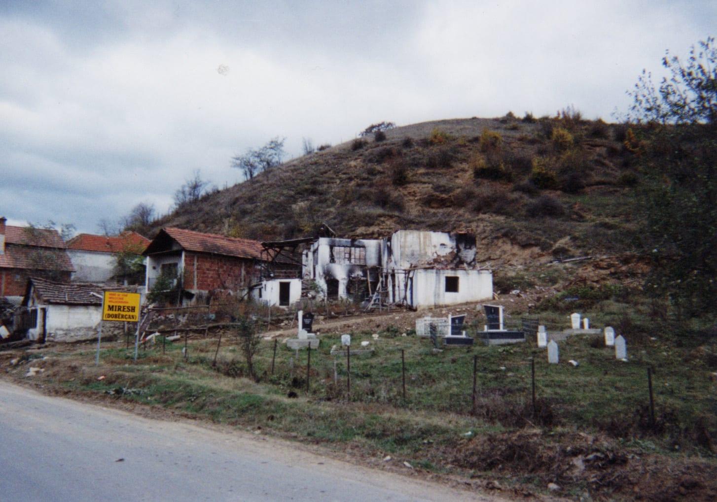 Burnt-out roofless white houses at the foot of a hill beside a road and a village cemetery.