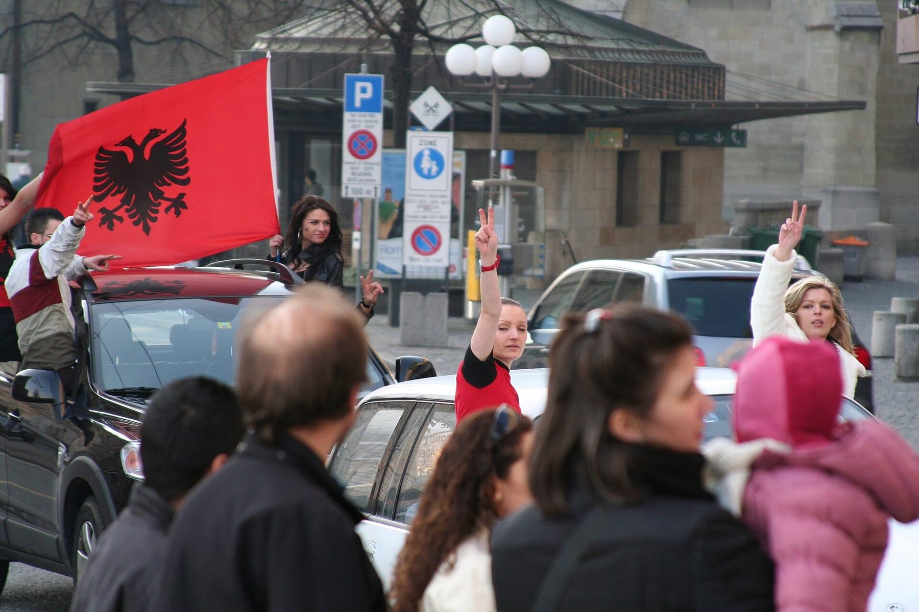 Young people wave the Albanian flag from car windows; passers-by greet them.