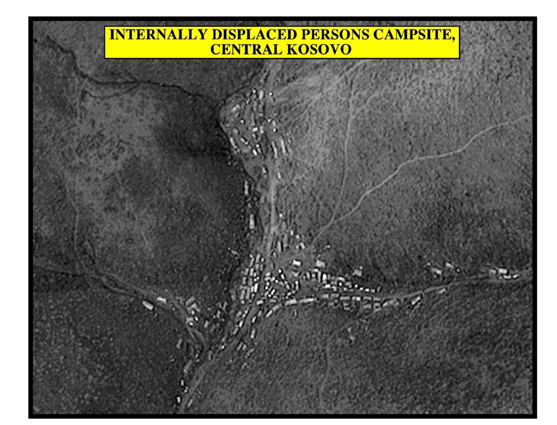 Black-and-white aerial view of a valley with vehicles and makeshift shelters along tracks; caption 'Internally displaced persons campsite, Central Kosovo'.
