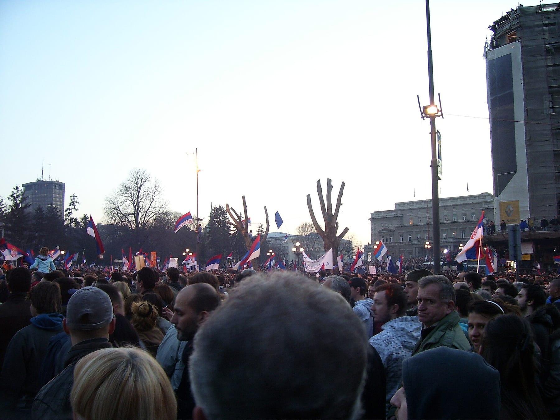 Large crowd with Serbian flags in a square at dusk.