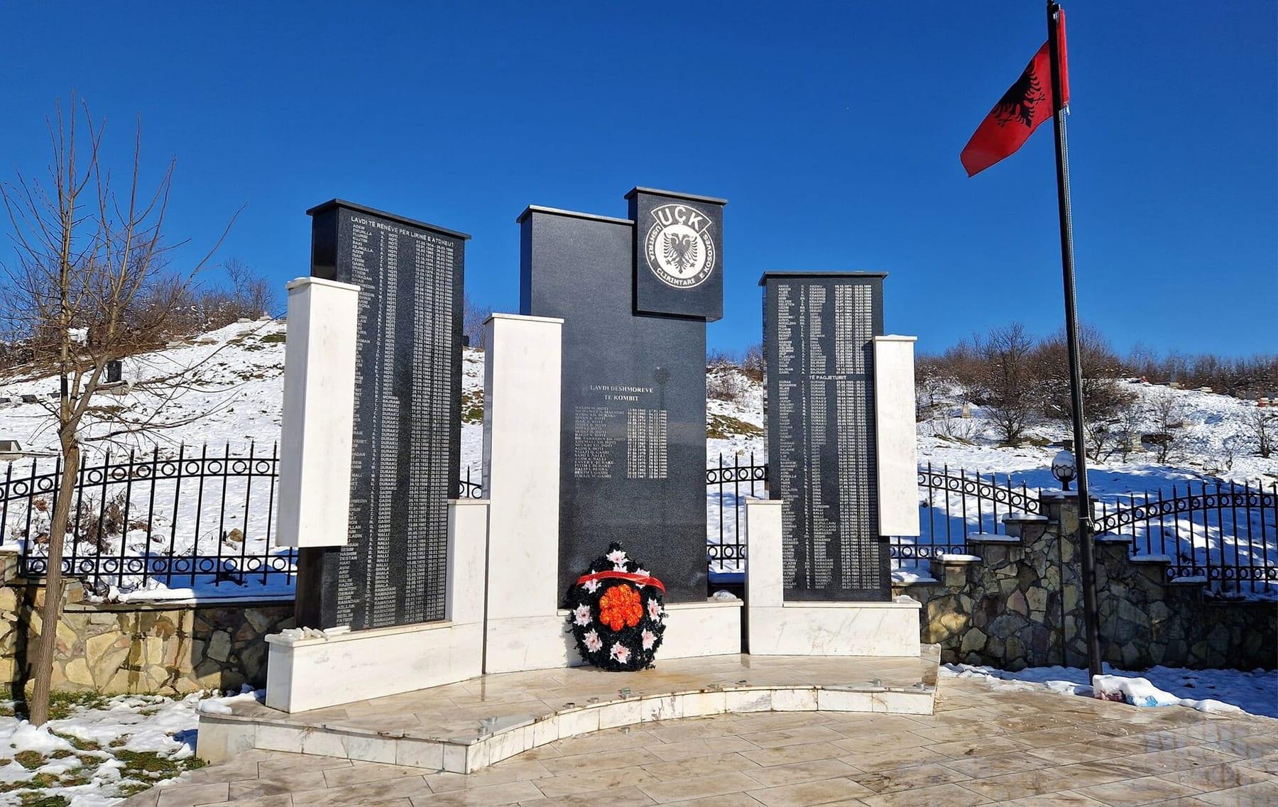 Black stone slabs with names, a wreath and an Albanian flag at a snow-covered memorial site.