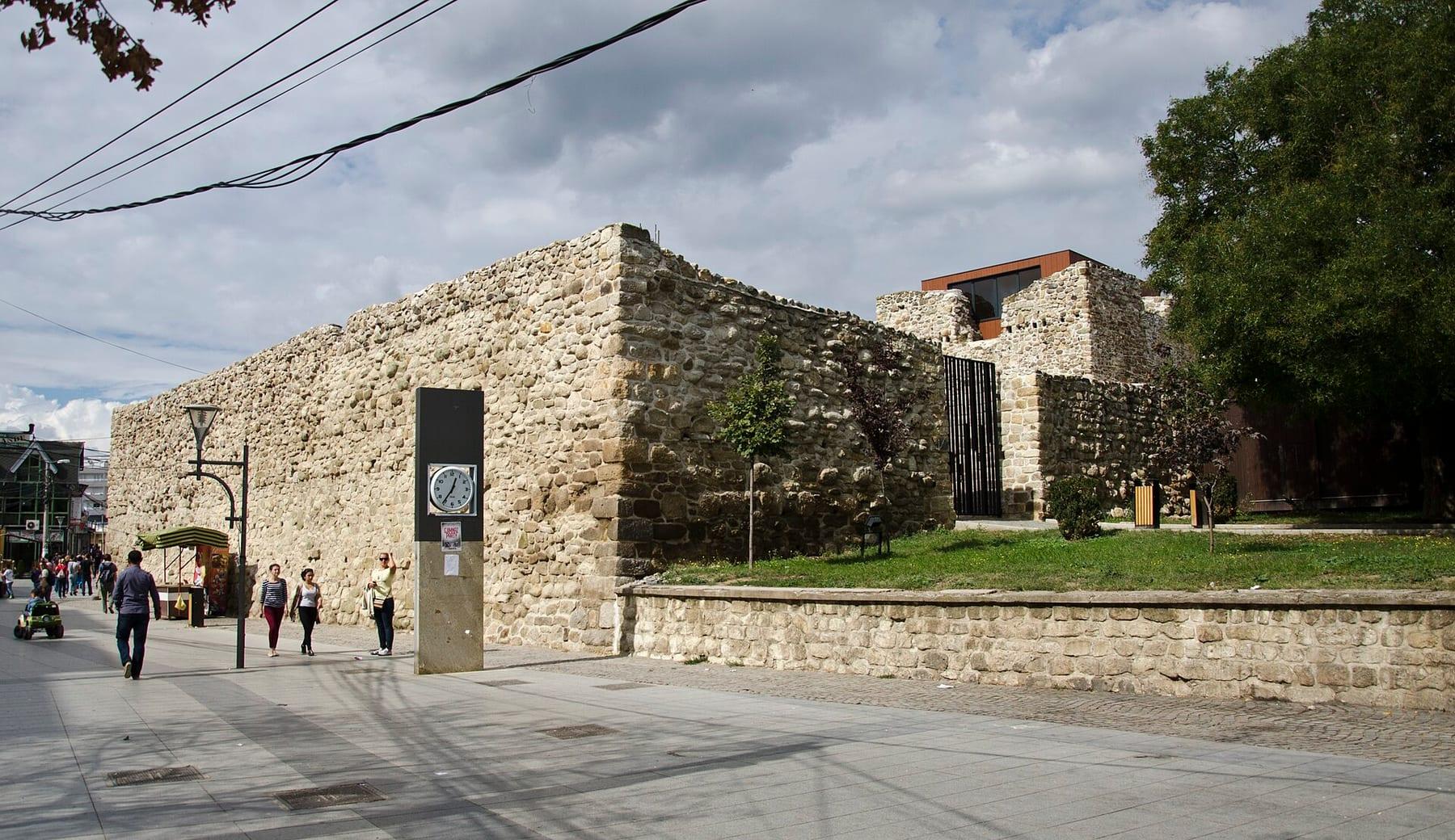 High stone walls of the fortress in the town centre with passers-by in the square.