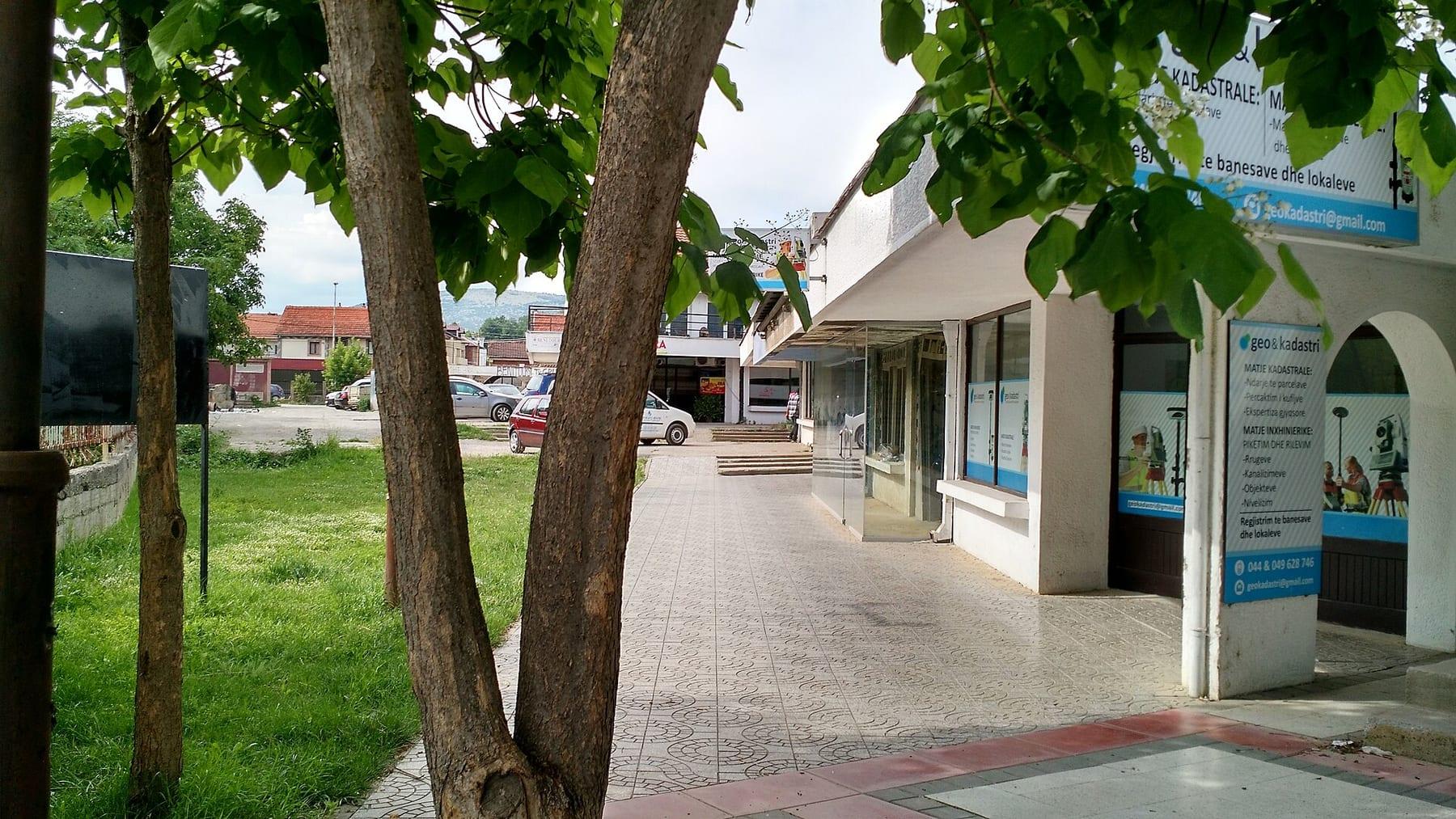 Paved sidewalk with trees and shopfronts of a small shopping centre.