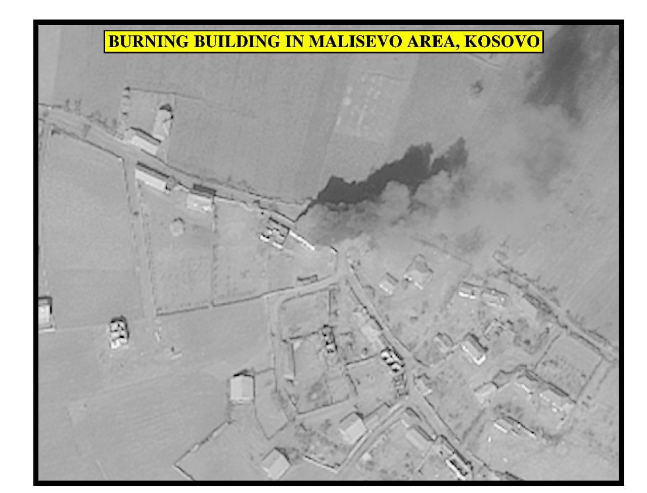 Black-and-white aerial view of a village with a plume of smoke rising from a building.