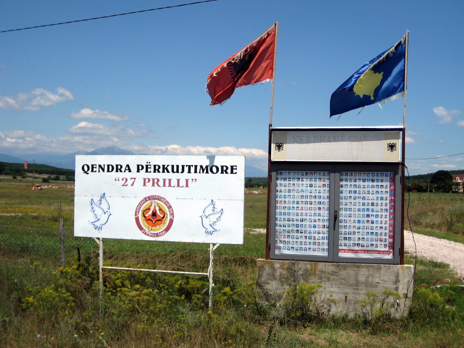 Sign reading 'Qendra Përkujtimore 27 Prilli' and a display case of small photographs, with the Albanian and Kosovo flags.