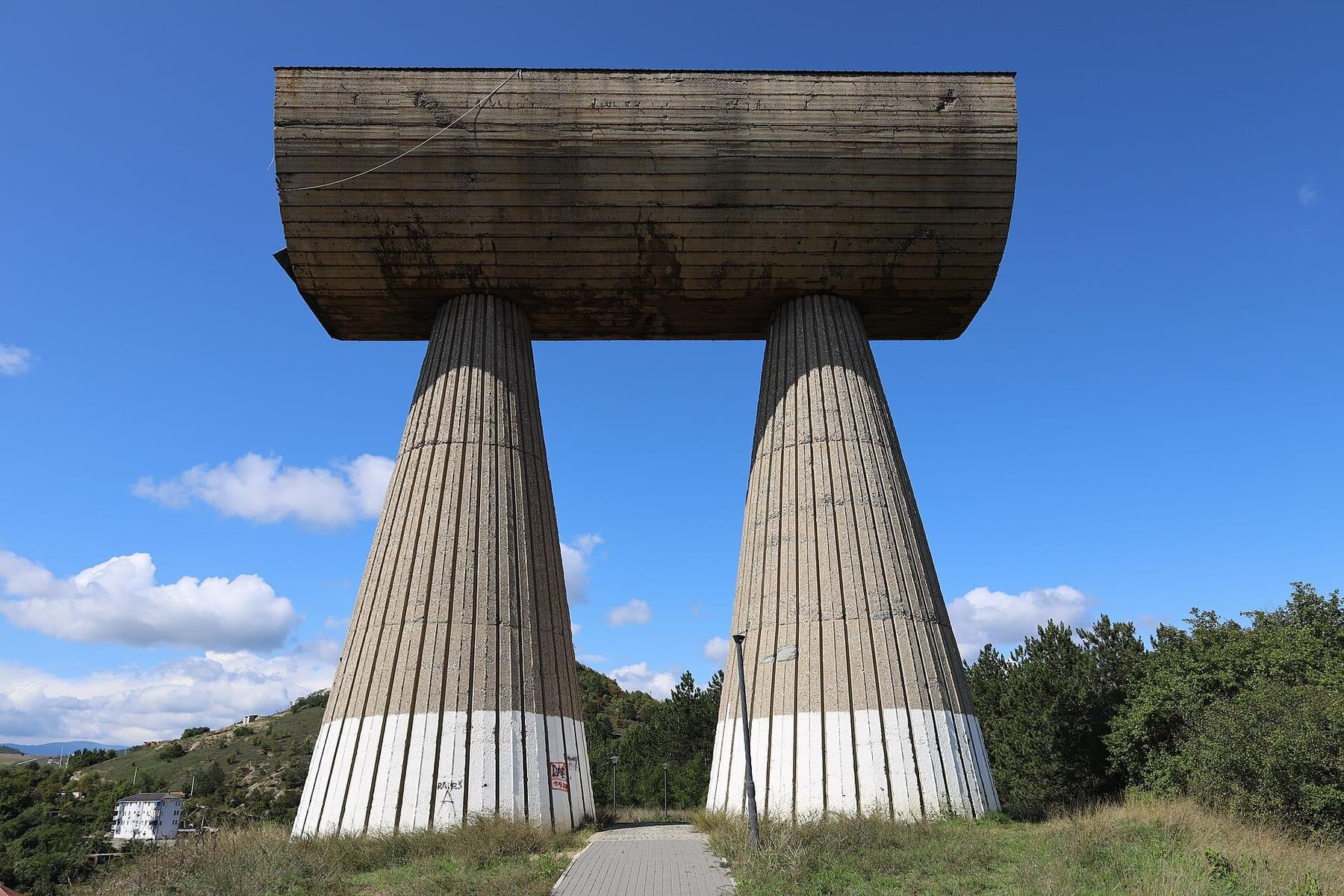 Two conical concrete columns carrying a horizontal trough, on a hill under a blue sky.