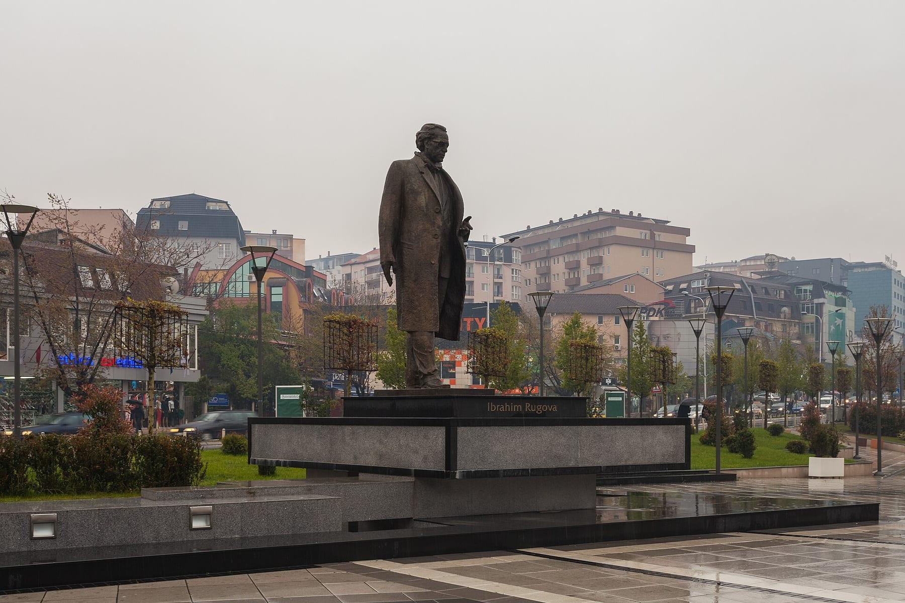 Bronze statue of a standing man on a dark base, with city buildings behind.
